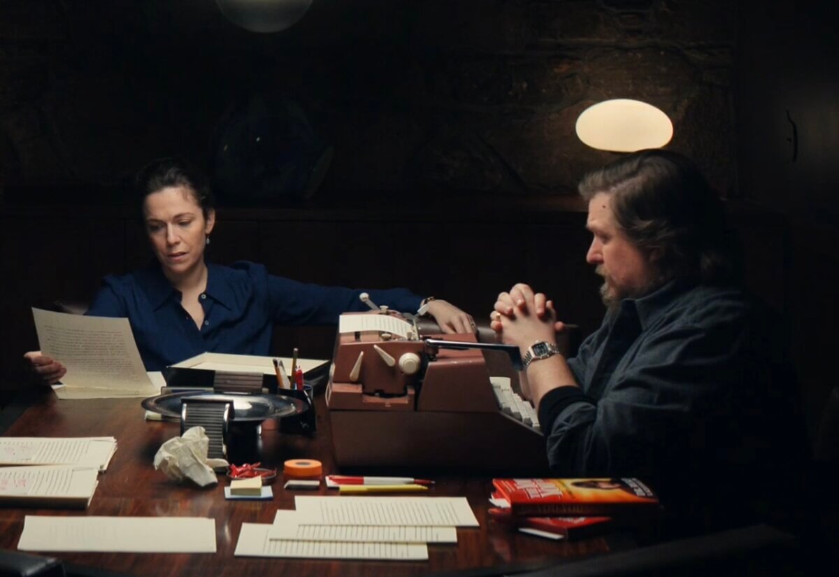 A scene from Severance. Ricken and Devin sit at a darkly-lit dining table, its surface covered with neatly arranged typed pages, paper-clipped stacks of paper, a crumpled page, etc. Ricken has his hands clapsed in front of him, and beneath his hands is the biggest, brownest typewriter the production designers could find. It seems to be an Optima M16